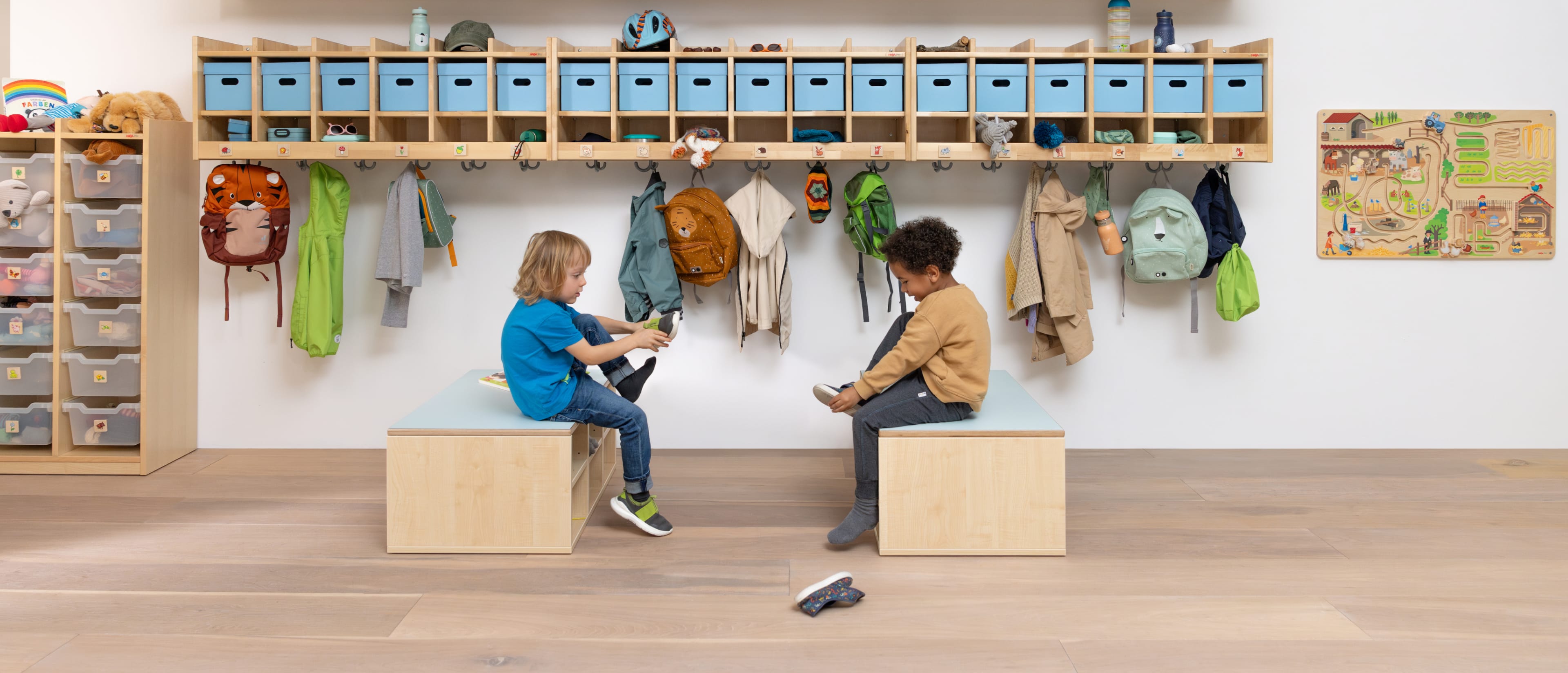 Zwei Kinder sitzen vor einer Kita-Garderobe auf Gardeorbenbänken und ziehen sich ihre Schuhe an.