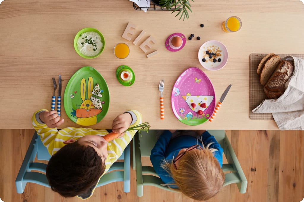 Osterrituale mit Kindern |  Zwei Kinder sitzen am Tisch, vor Tellern in Ei-Form mit Ostermotiven geschmückt