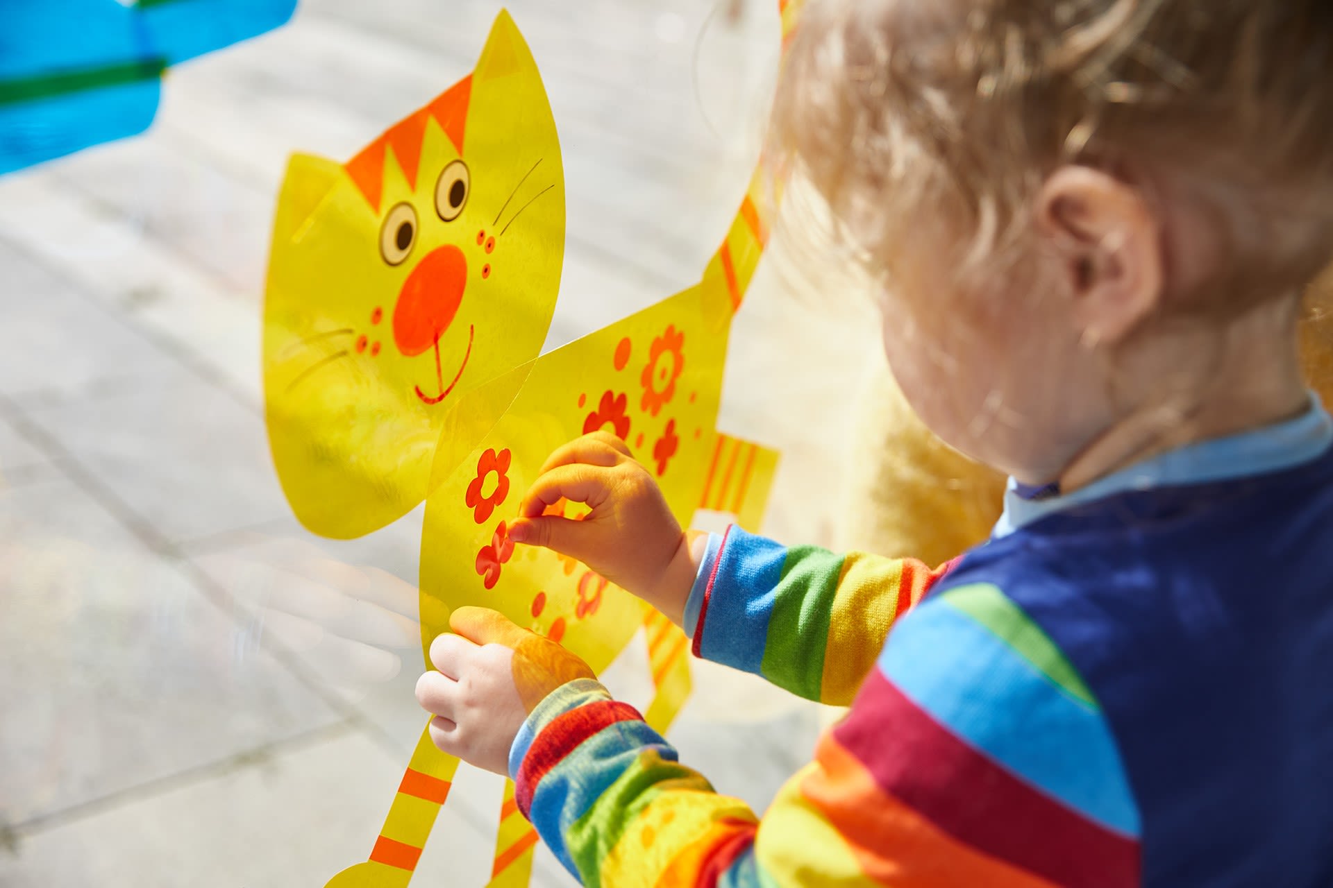 Basteln mit Kindern |  Mädchen klebt Fensterbild, gelbe Katze mit roten Blumen an Fensterscheibe