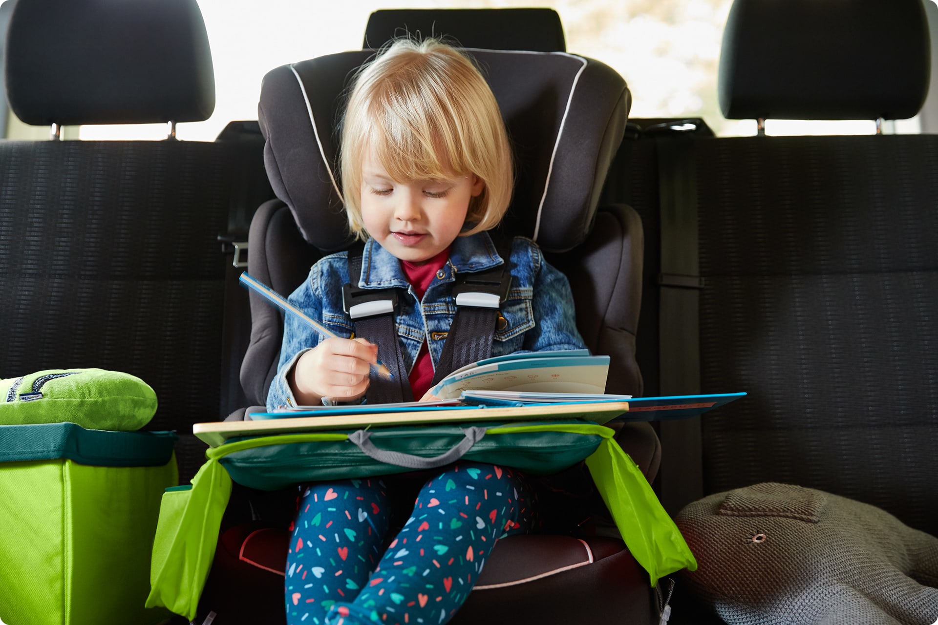 Kinderspiele im Auto | Kleines Mädchen angeschnallt im Autositz mit Maltisch-Kissen auf dem Schoß malt