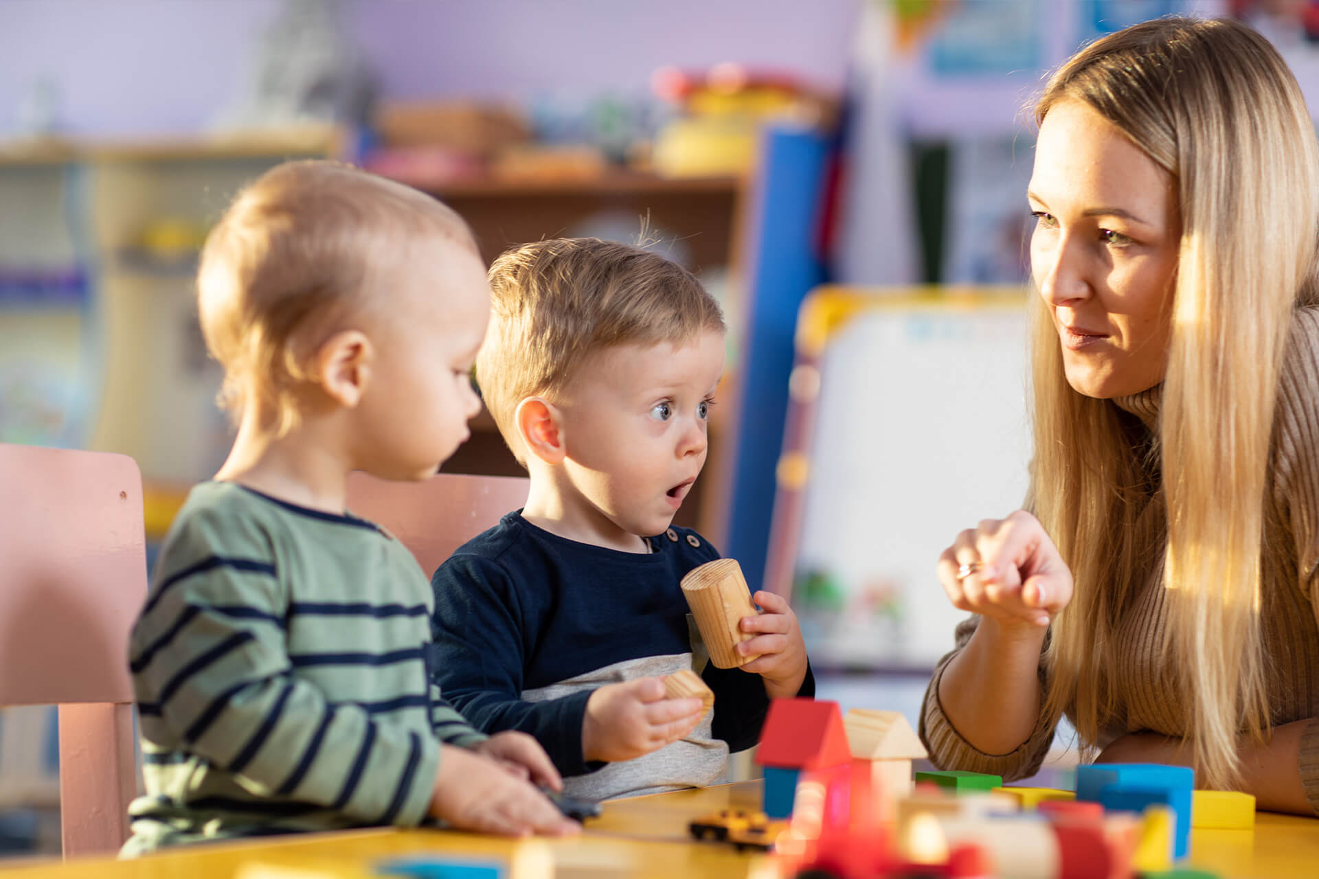 Grid | 3 | Education | Image| Krippe | 2 Kinder spielen mit Bauklötze, Erzieherin sitzt daneben