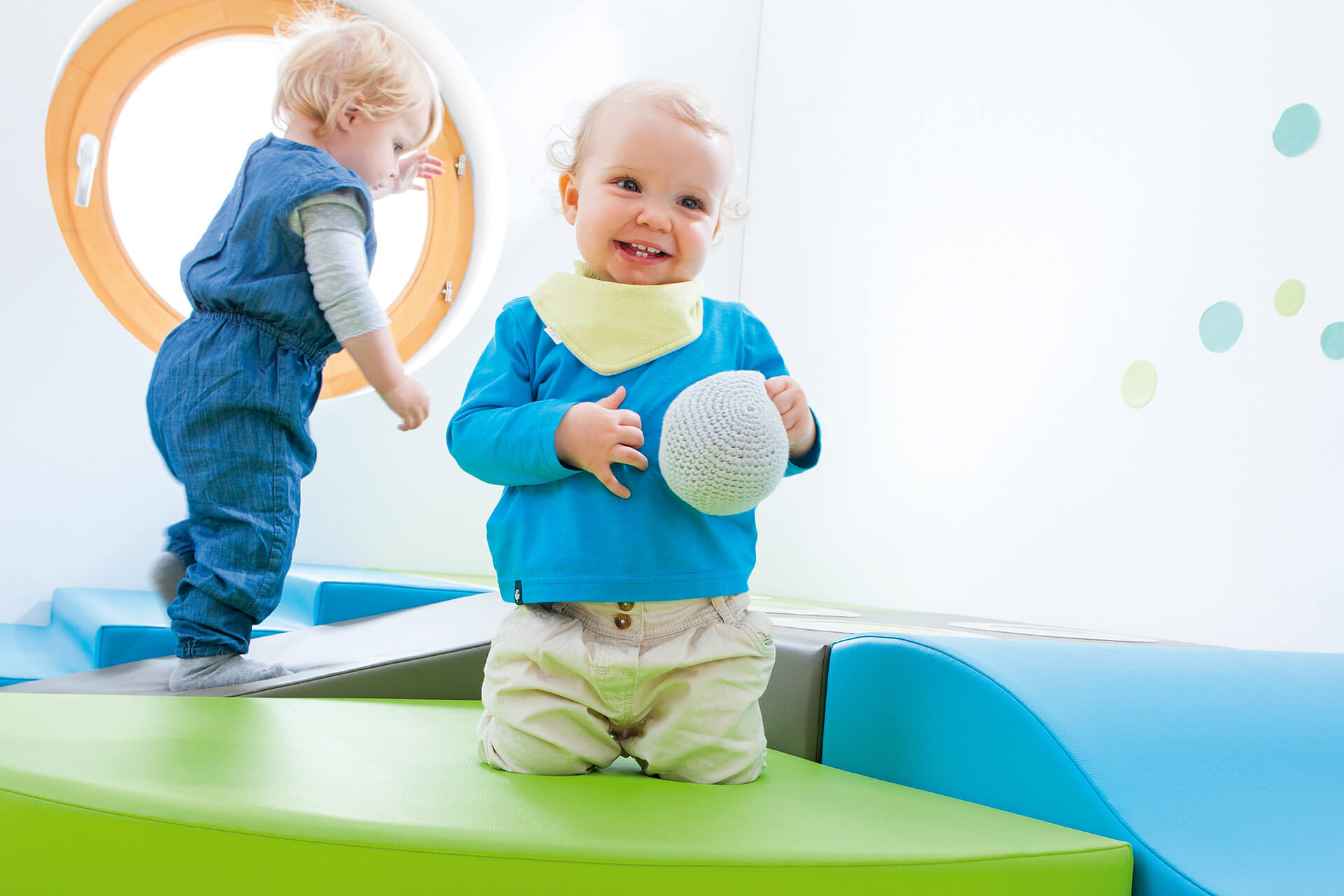 Education | Krippe | 2 | Image | Spielen & Bewegung | kleines Kind hat einen Schaumstoffball in der Hand und kniet auf einem grünen Schaumstoffpodest