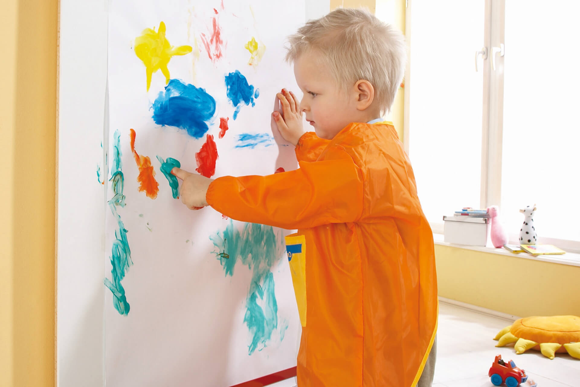 Education | Krippe | 2 | Image | Kreativ- & Bastelbedarf | kleiner Junge hat orangefarbenen Malkittel an und malt mit Finger an einer Wand