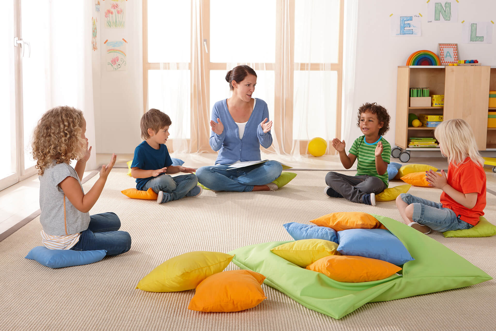 Ruheraum | 2 | Kissen & Decken | Erzieherin und Kinder sitzen auf bunten Kissen im Kreis und singen und klatschen