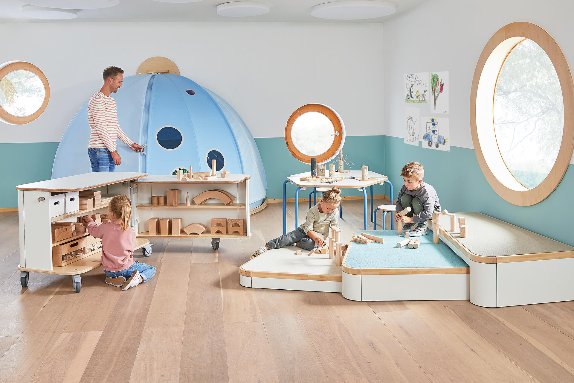 Spielraum | Erzieher mit drei Kindern, die auf einem Dreier-Podest mit Bauklötzchen spielen, ein Mädchen sortiert Spielzeug in den Materialschrank, Erzieher steht am hellblauen Kuppelzelt