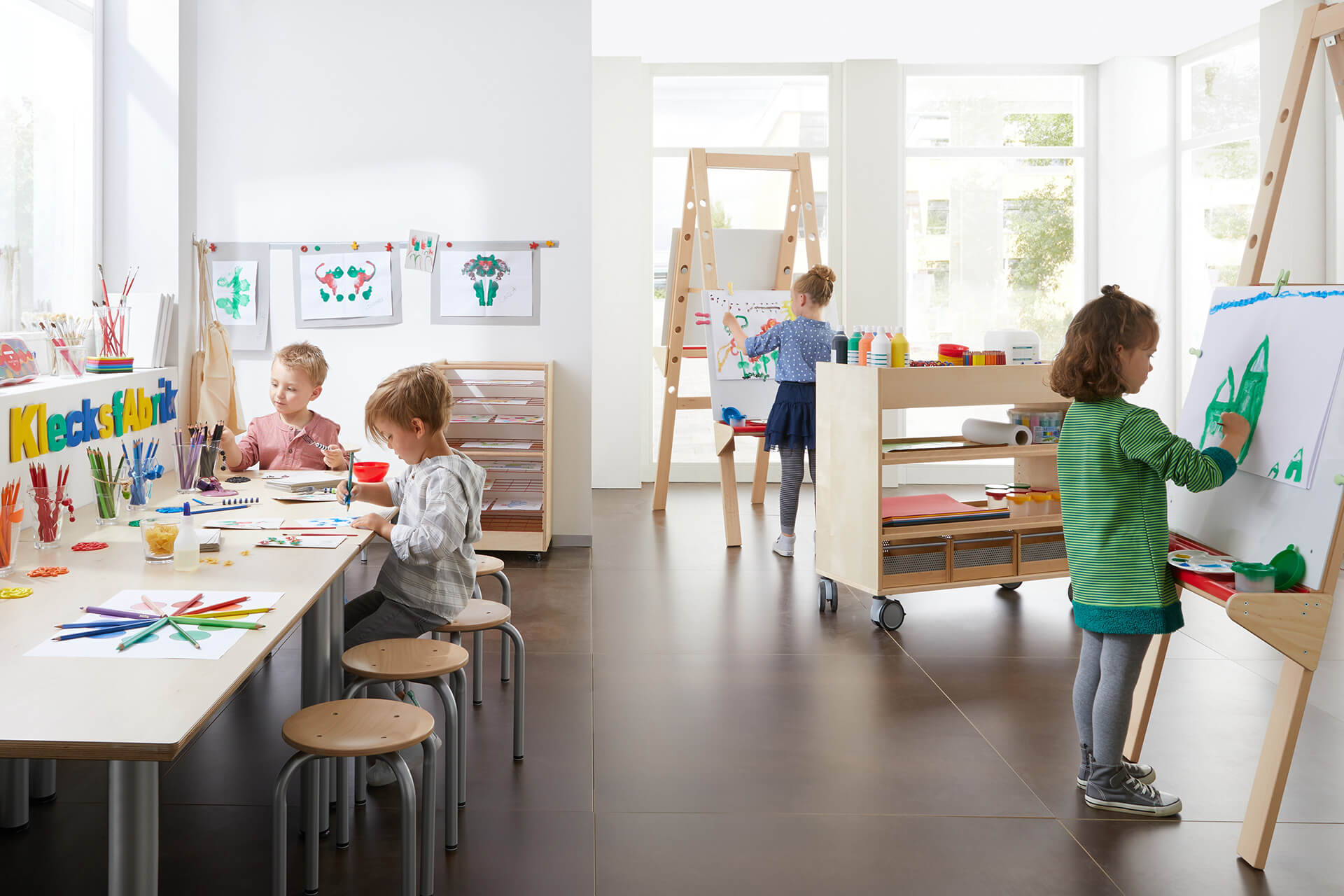 Spielraum | Kreativ- & Bastelbedarf | Zwei Mädchen malen Bilder an einer Staffelei mit Buntstiften, zwei Kinder sitzen auf Hockern und malen Bilder am Tisch