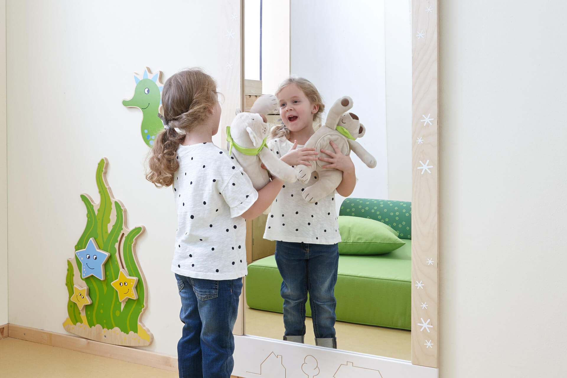 Räume | Krippe & Kindergarten | Wasch- und Wickelraum | Blondes Mädchen mit Zopf und gepunktetem T-Shirt, steht vor einem Spiegel, mit Teddybär im Arm