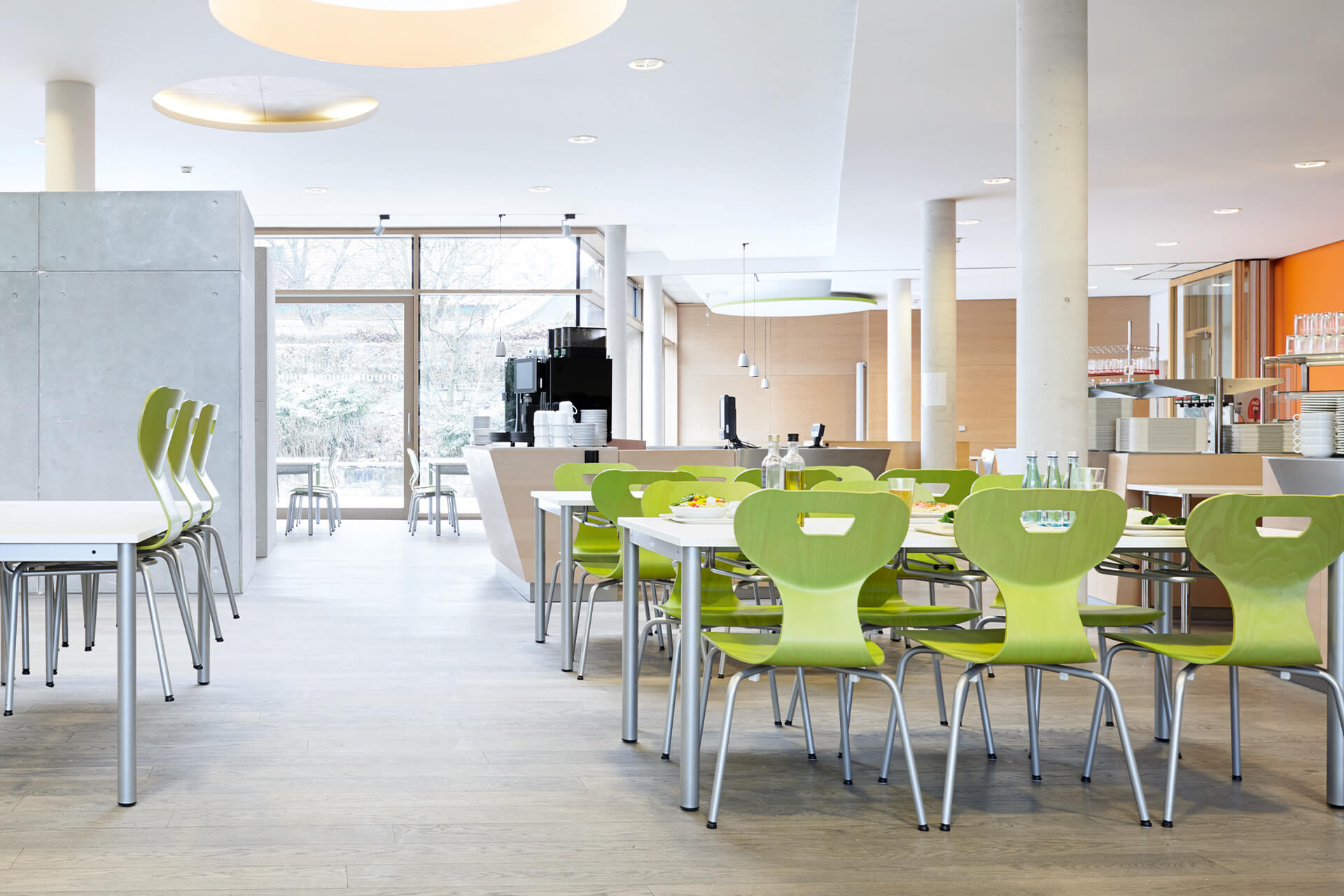 Schulcafeteria | Cafeteria-Raum eingerichtet mit Tischen und grünen Stühlen, mit Theke und großen  Bodenfenstern