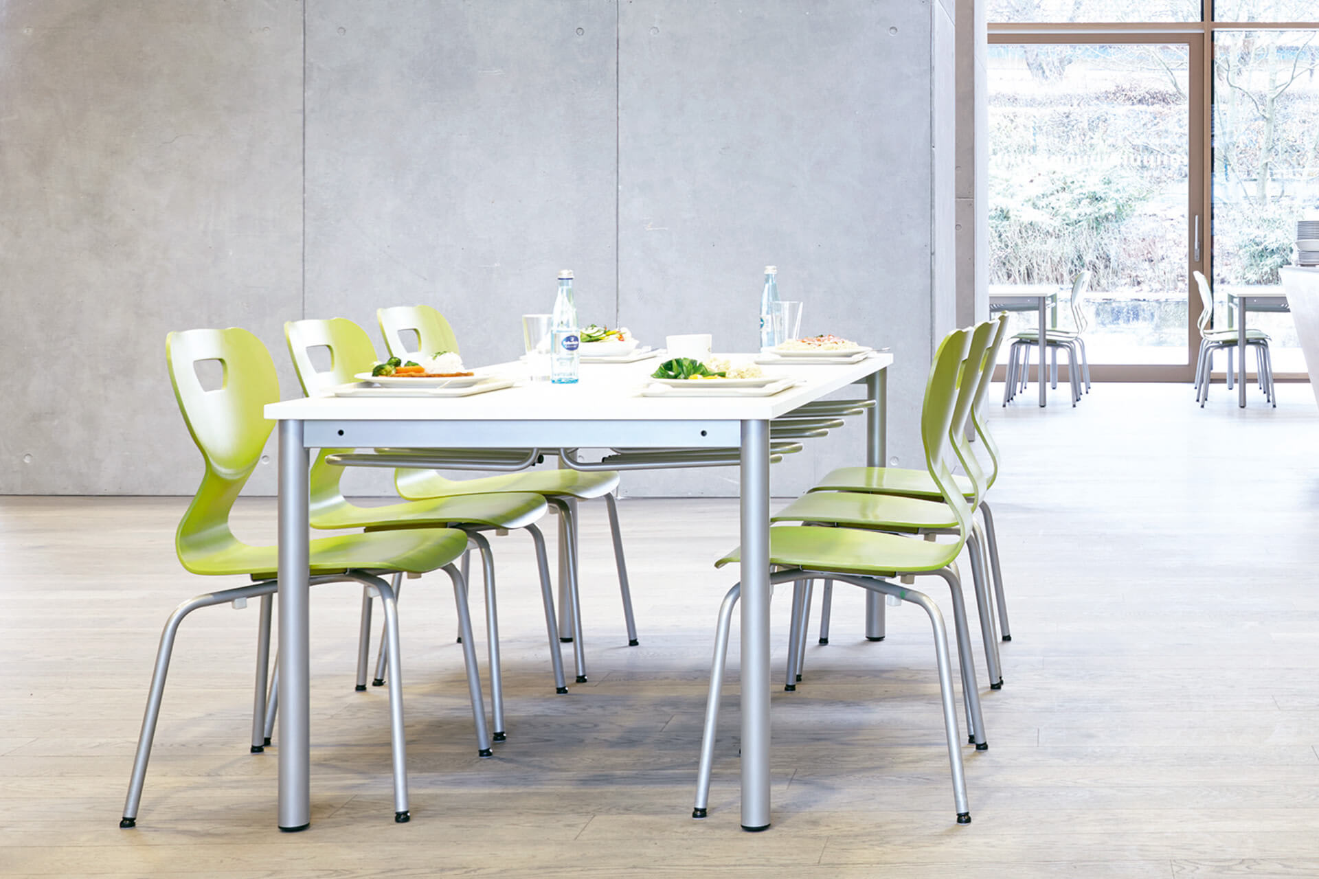 Schulcafeteria | Gedeckter Tisch mit Tellern, Gläsern und Flaschen, an dem sechs Stühle mit grünem Sitzteil, steht vor einer Betonwand