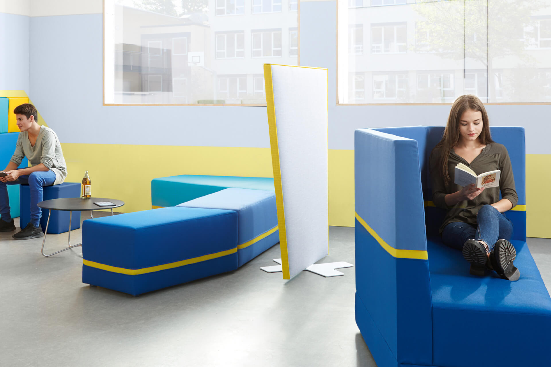 Schulcafeteria | Raumteiler | Mädchen sitzt auf einem blauen Sofa und liest, daneben eine Sitzgruppe, getrennt durch einen graue Trennwand