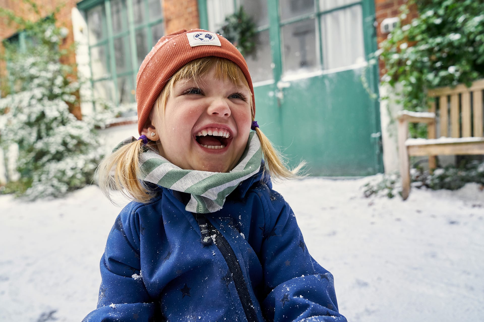  Materialinfo Softshell | Lachendes Mädchen mit brauner Mütze, grün-gestreiften Schal und blauer Softshelljacke sitzt im Schnee