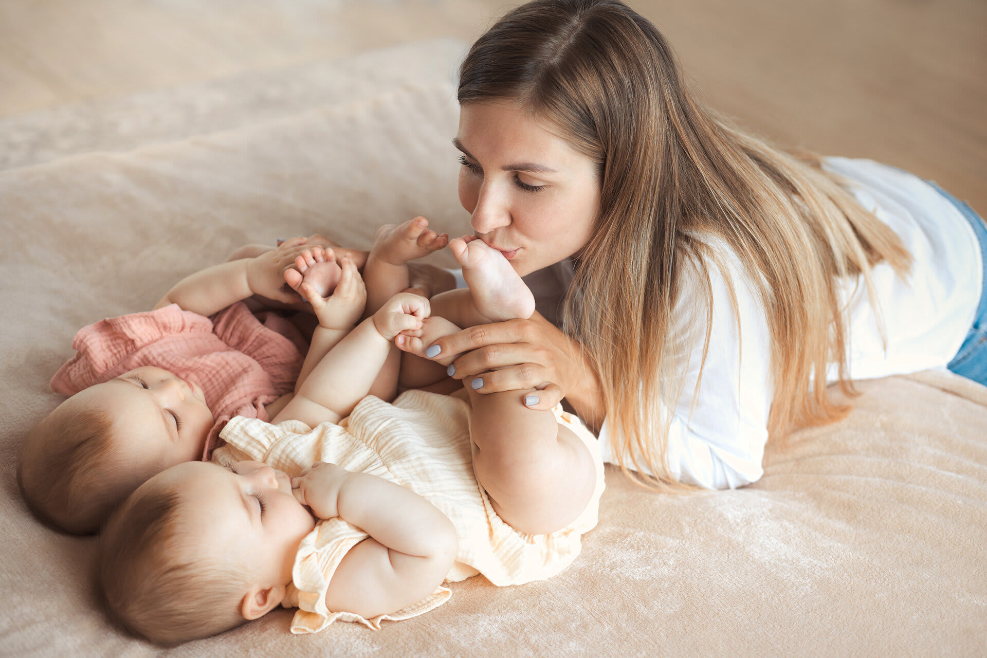 Rituale für Babys & Kleinkinder | Mutter küsst barfuß Beine von zwei Baby-Zwillingsschwestern auf dem weichen Bett zu Hause
