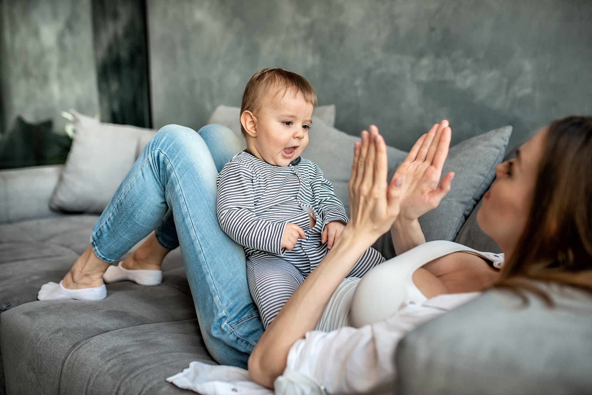 Rituale für Babys & Kleinkinder | Mutter sitzt mit angewinkelten Beinen auf dem Sofa, Baby liegt auf den Beinen und Mutter klatscht in die Hände