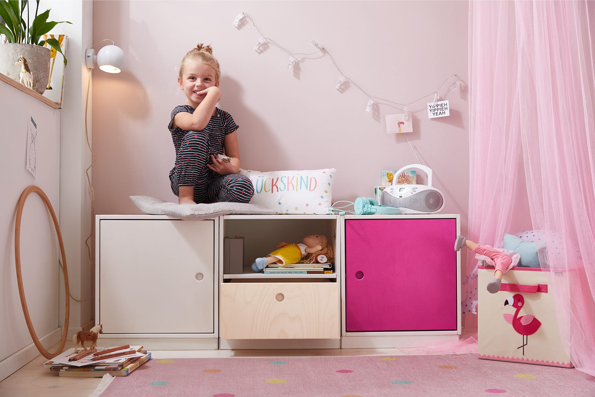 Kinderzimmer einrichten | Mädchen sitzt auf Regalsystem mit weißer und pinker Schranktür