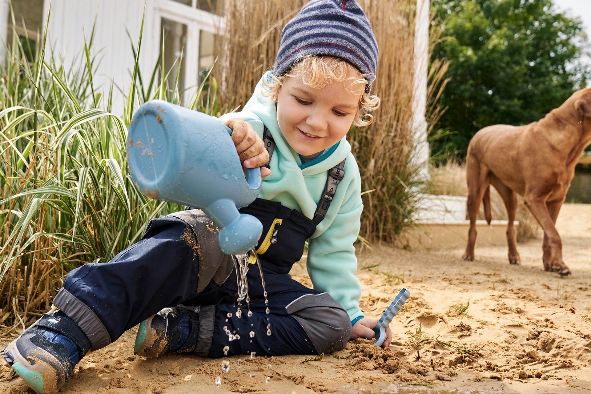 Spiele für draußen | Kind mit Matschhose, Fleeceshirt und Mütze, sitzt im Sand, hat einen Wassergießer in der Hand.