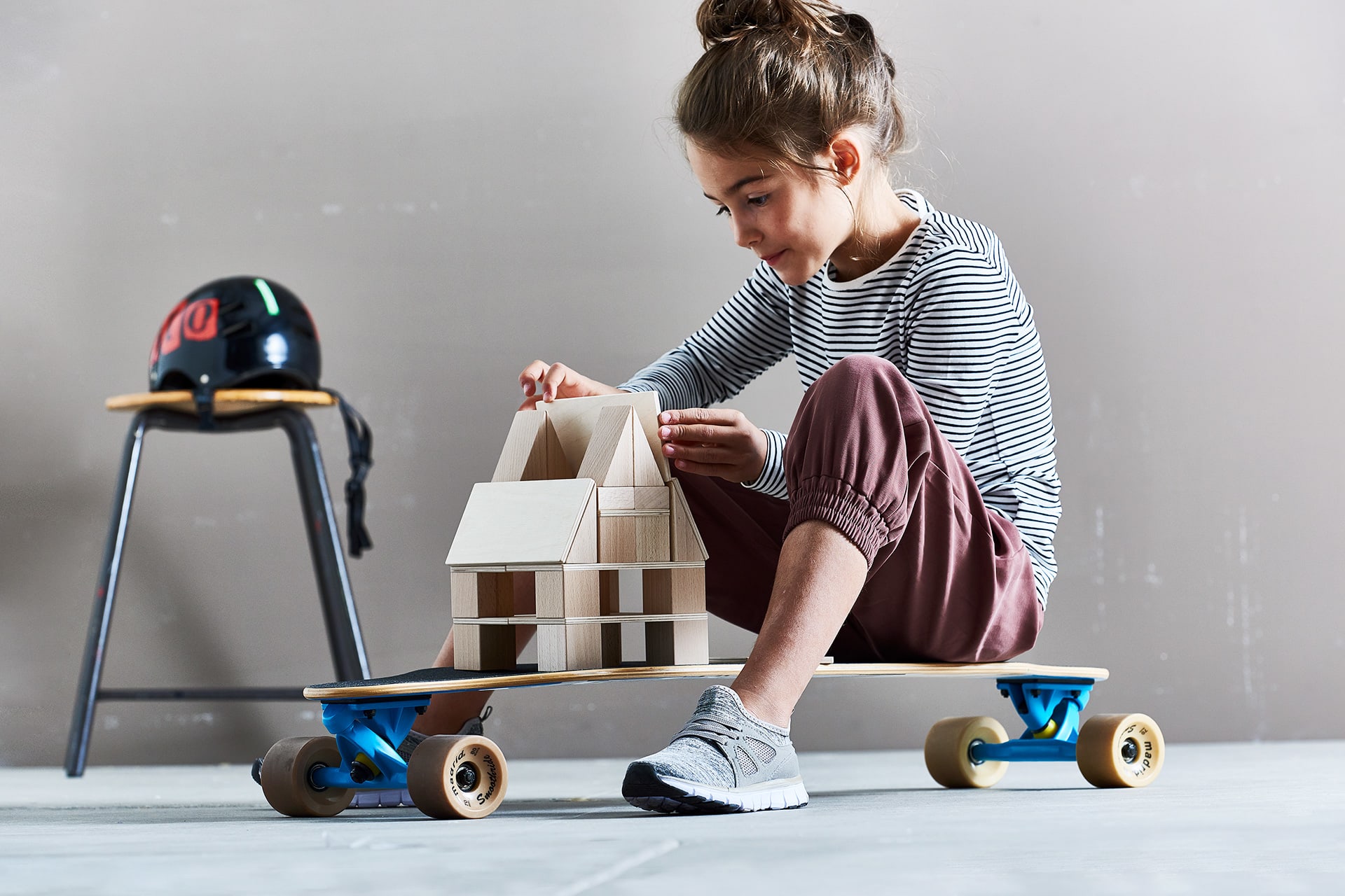Bauen | Mädchen mit braunen Haaren baut auf Skateboard ein Haus mit Holzbausteinen