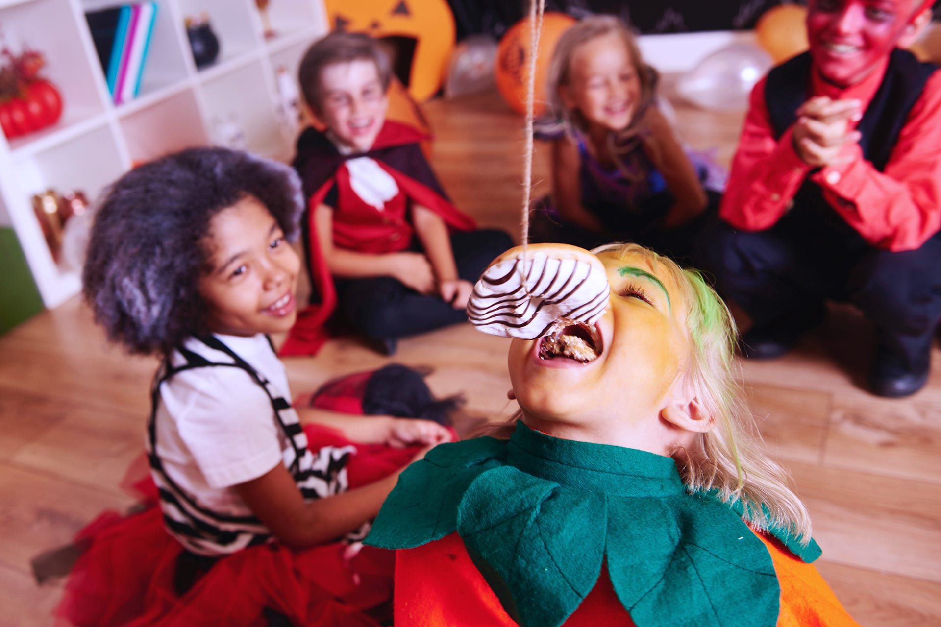 Halloween Spiele | Verkleidetes Mädchen beißt in einen aufgehängten Donut, sitzt in der Gruppe