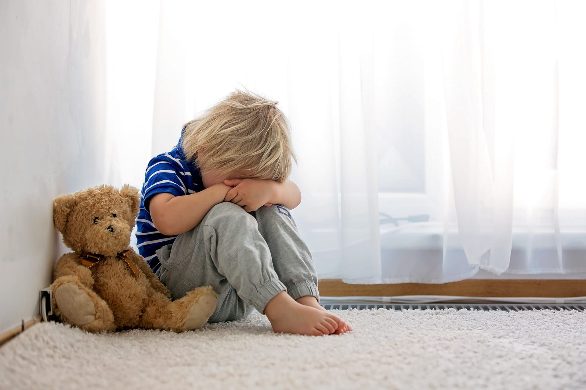 Auf Kinder eingehen | Kind sitzt mit Teddy allein im Zimmer auf einem weißen Teppich