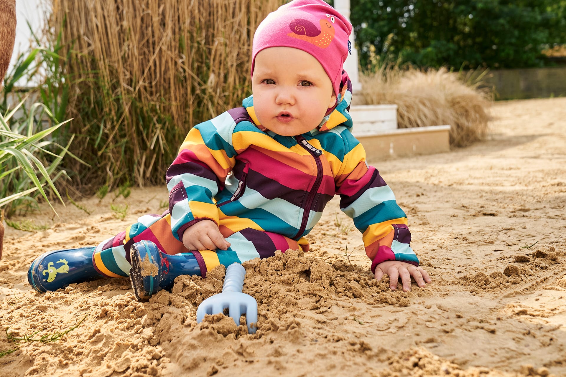 Kita Gebühren, Kindergarten Kosten | Kleinkind sitzt mit geringeltem Softshell-Anzug im Sand