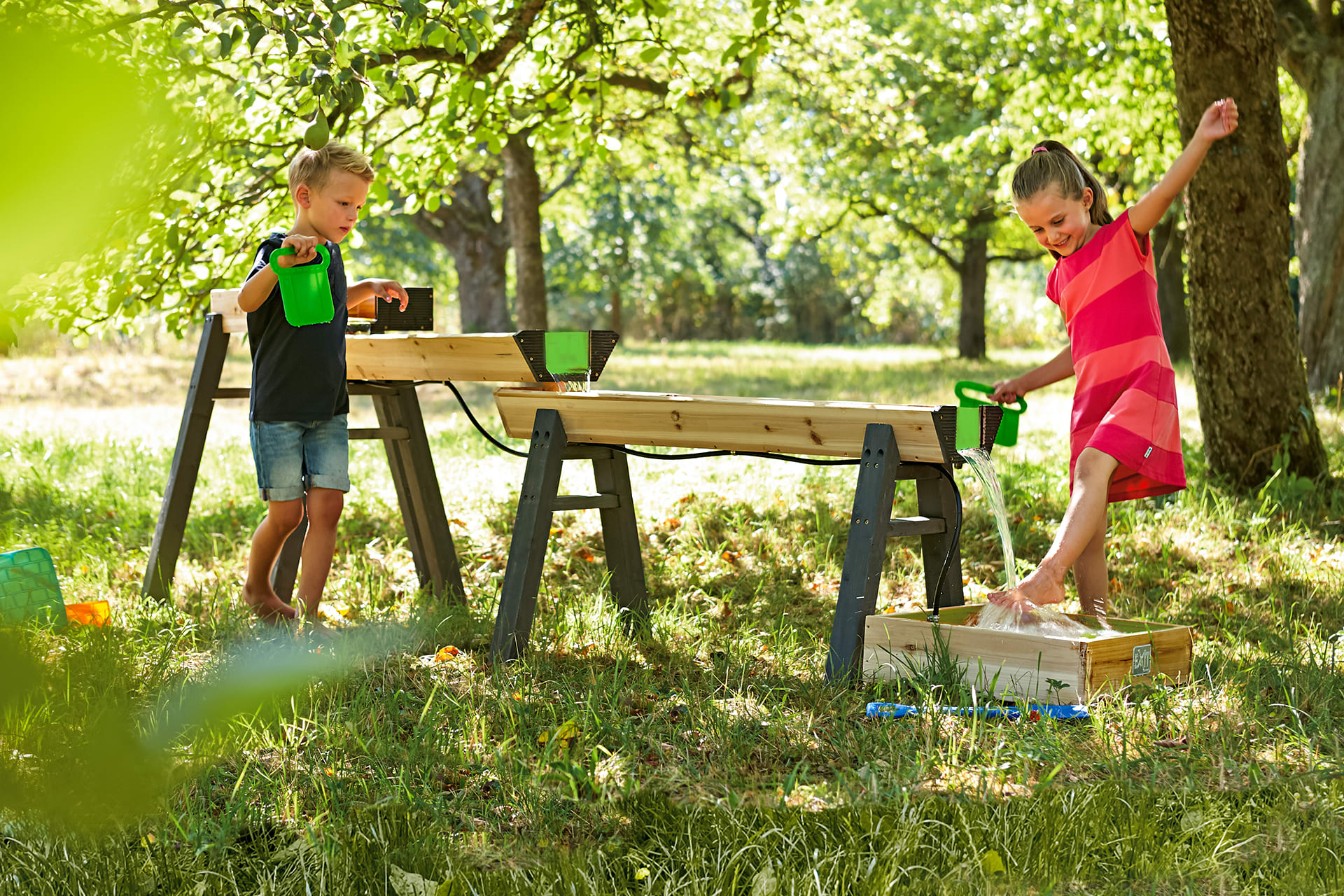 Wasserspiele draußen | Mädchen im roten Kleid spielt mit Jungen an Wasserspielbahn aus Holz