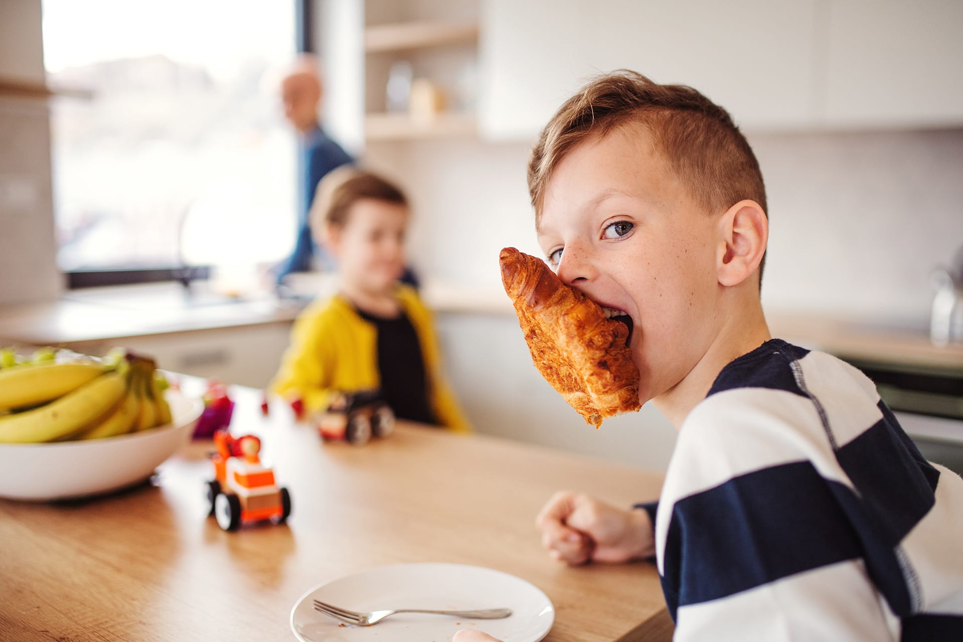 Kindern Tischmanieren beibringen | Junge hat Butterhörnchen am Mund, sitzt am Esstisch
