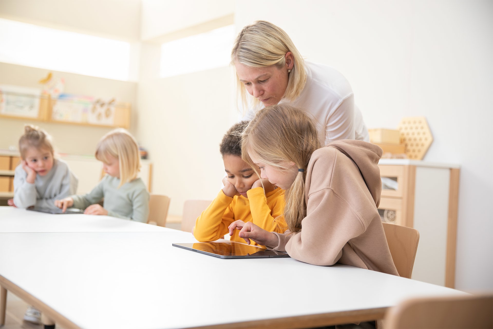 Medienkompetenz | 2 | Image | Tablet als Lerninstrument | Erzieherin schaut über die Schulter von 2 Kinder die mit Tablet lernen 