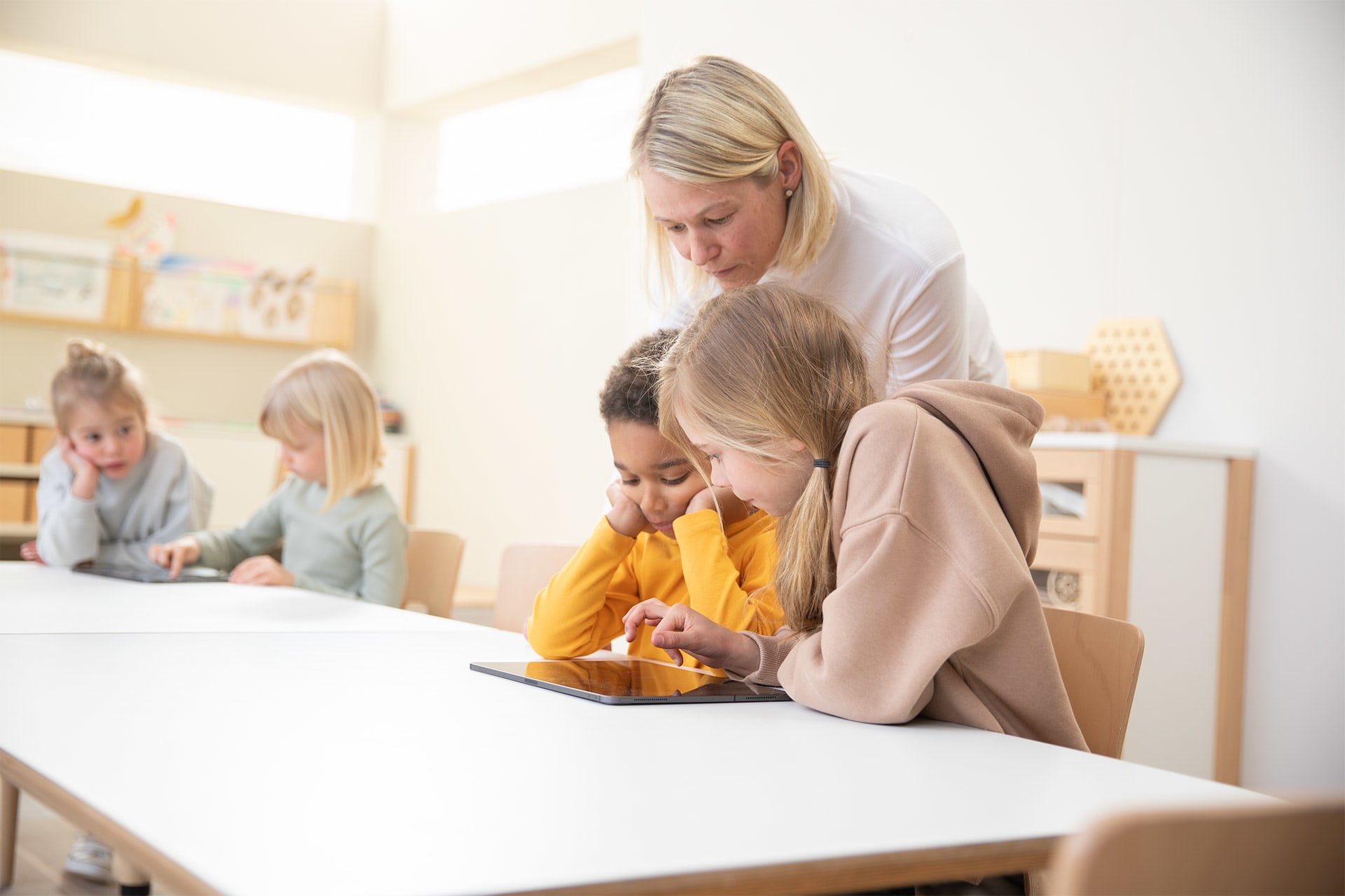 Medienkompetenz | 2 | Image | Tablet als Lerninstrument | Erzieherin schaut über die Schulter von 2 Kinder die mit Tablet lernen 