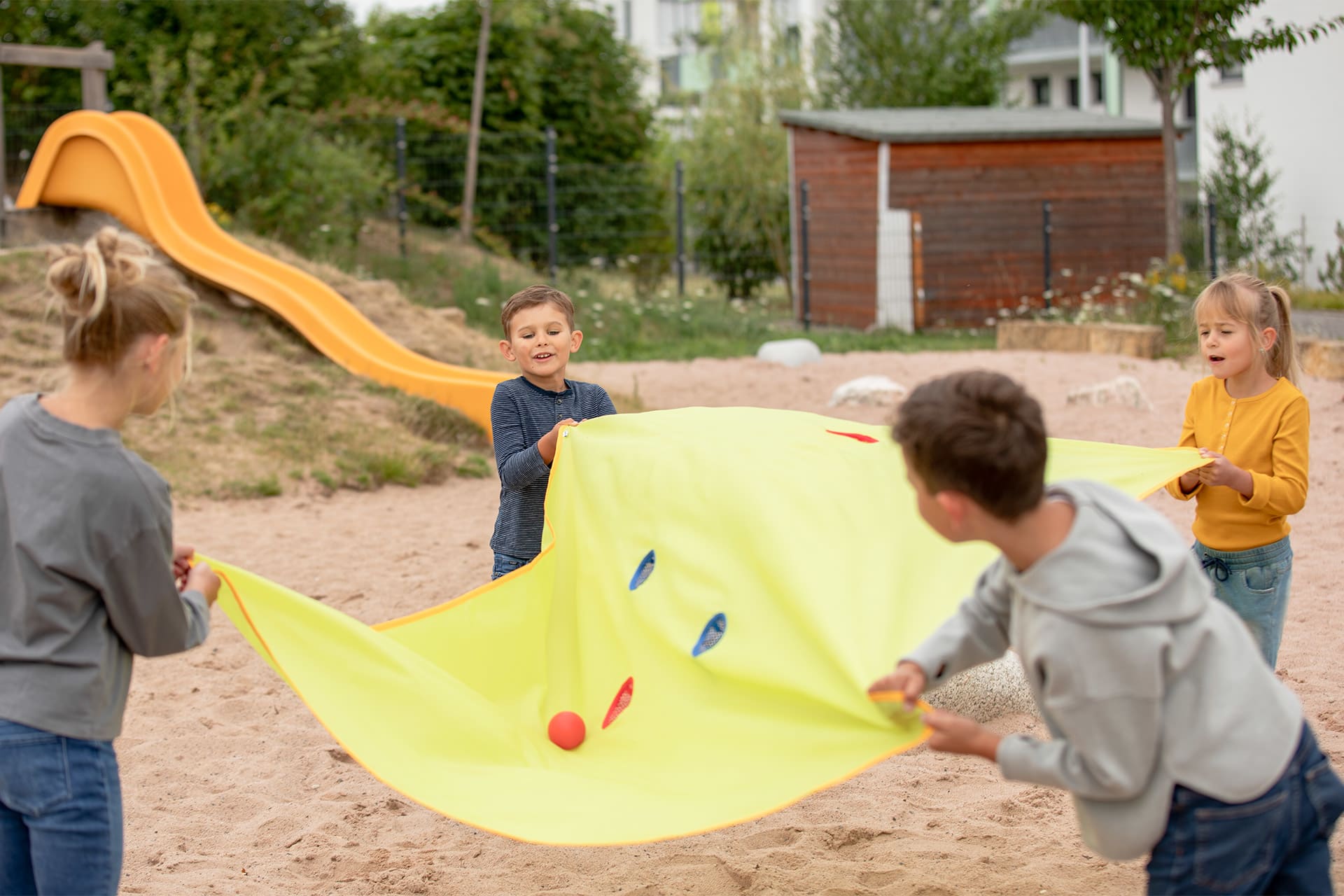Teamfähigkeit | 2 | Image | Tuchgolf | Kinder spielen mit gelben Tuchgolf auf einer Sandfläche. Gelbe Rutsche im Bildhintergrund