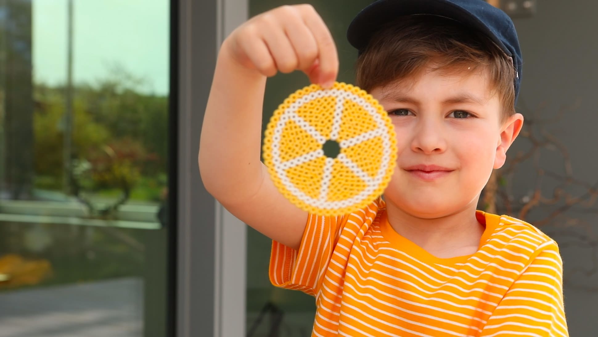 Ein Junge mit orangefarbenen T-Shirt hält einen Wespenschutz aus Bügelberlen in Orangenformen hoch.