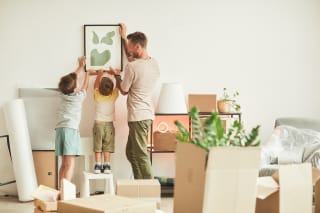  Umzug mit Kindern | Ganzseitiges Porträt eines glücklichen Vaters mit zwei Söhnen, der beim Einzug in ein neues Haus Bilder an die Wand hängt