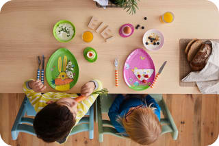 Osterrituale mit Kindern |  Zwei Kinder sitzen am Tisch, vor Tellern in Ei-Form mit Ostermotiven geschmückt