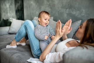 Rituale für Babys & Kleinkinder | Mutter sitzt mit angewinkelten Beinen auf dem Sofa, Baby liegt auf den Beinen und Mutter klatscht in die Hände