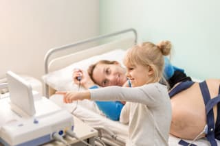 Schwangerschaft 40 SSW | Schwangere Frau mit Tochter lächelnd und Blick auf das Display während der Schwangerschaftsuntersuchung im Krankenhaus