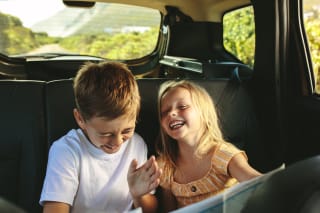  Kinderspiele selber machen | Kleines Mädchen und Junge sitzen im Auto und lachen, vor einem Buch