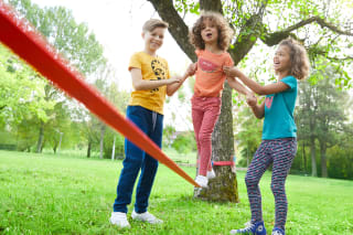 Sport mit Kindern | Mädchen balanciert auf Slackline und wird auf beiden Seiten von Kindern gestützt