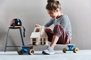 Bauen | Mädchen mit braunen Haaren baut auf Skateboard ein Haus mit Holzbausteinen