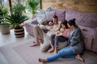Geschichten spannend erzählen | Zwei glückliche Schwestern mit Mutter und Vater sitzen auf dem Boden, umarmen sich und lesen ein Buch zu Hause