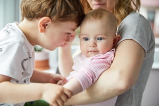 Familienzusammenhalt | Mutter hat kleines Baby auf Arm und Geschwisterkind sucht Nähe