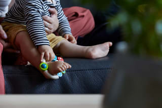  Spielerisch lernen |  Baby sitzt auf Mamas Schoß, hat Greifling um den Fuß gelegt