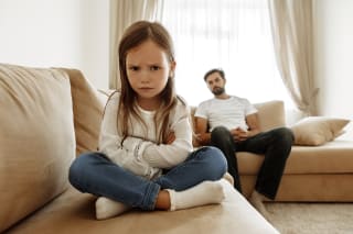 Trotzphase bei Kindern | Mädchen im Schneidersitz auf dem Sofa, hat die Arme verschränkt und schaut bockig, Papa sitzt dahinter