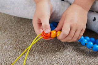 Fröbel - Prickeln, Sticken, Fädeln | Kinderhand fädelt Perle auf eine gelbe Schnur