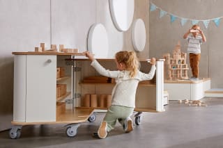 Spielraum | Möbel & Raumgestaltung | Mädchen kniet vor Material-Schrank und öffnet die Türen, daneben steht Junge auf Podest und baut einen Turm aus Holzbauklötzchen