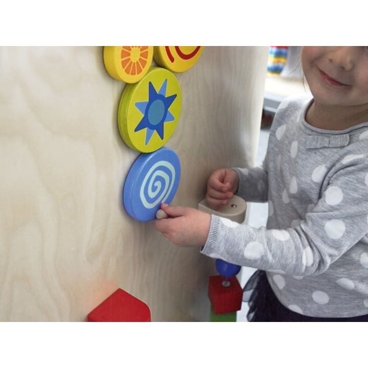 Ein Kind in einem grauen Polka-Dot-Shirt interagiert mit einer vertikalen Holztafel, auf der bunte kreisförmige Teile mit verschiedenen Mustern zu sehen sind.