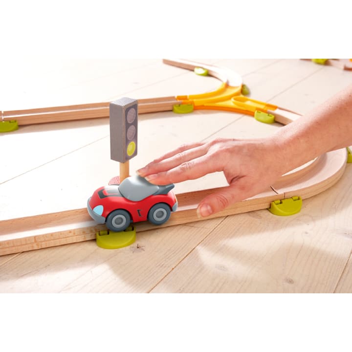 Eine Hand schiebt ein kleines rotes Spielzeugauto auf einer Holzeisenbahn, die an einer Spielzeugampel vorbeifährt. Die Bahn ist auf einem hellen Holzboden aufgebaut.