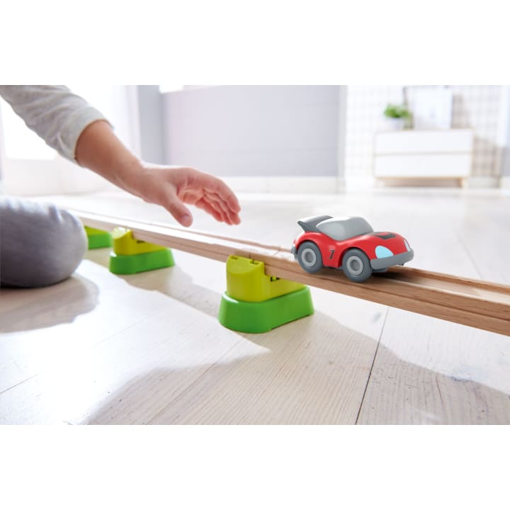 Eine Kinderhand greift nach einem kleinen roten Spielzeugauto auf einer hölzernen Schiene, die durch grüne Stützen erhöht ist, auf einem hellen Boden in einem hellen Innenraum.