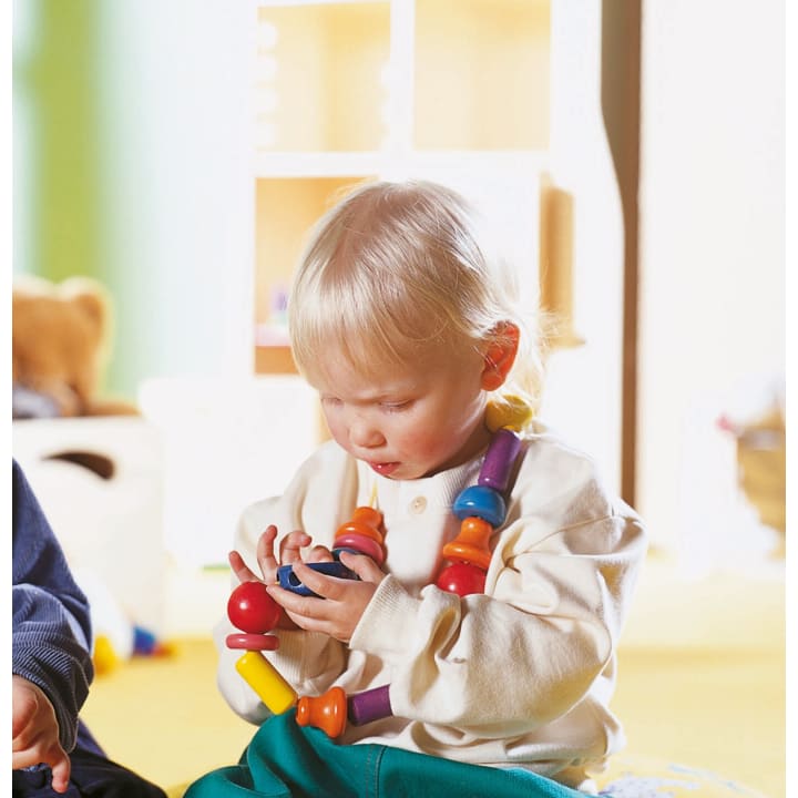 Ein kleines Kind mit hellem Haar, das ein cremefarbenes Hemd und eine Kette aus großen, bunten Perlen trägt, sitzt im Haus und konzentriert sich auf das Stapeln oder Untersuchen ähnlicher Spielzeugteile in seinen Händen.