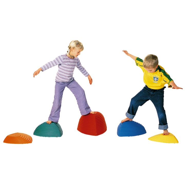 Zwei kleine Kinder balancieren auf bunten Plastik-Trittsteinen. Das Mädchen steht auf einem roten Stein, der Junge auf einem blauen. Sie strecken ihre Arme aus, um das Gleichgewicht zu halten, und scheinen miteinander zu spielen.
