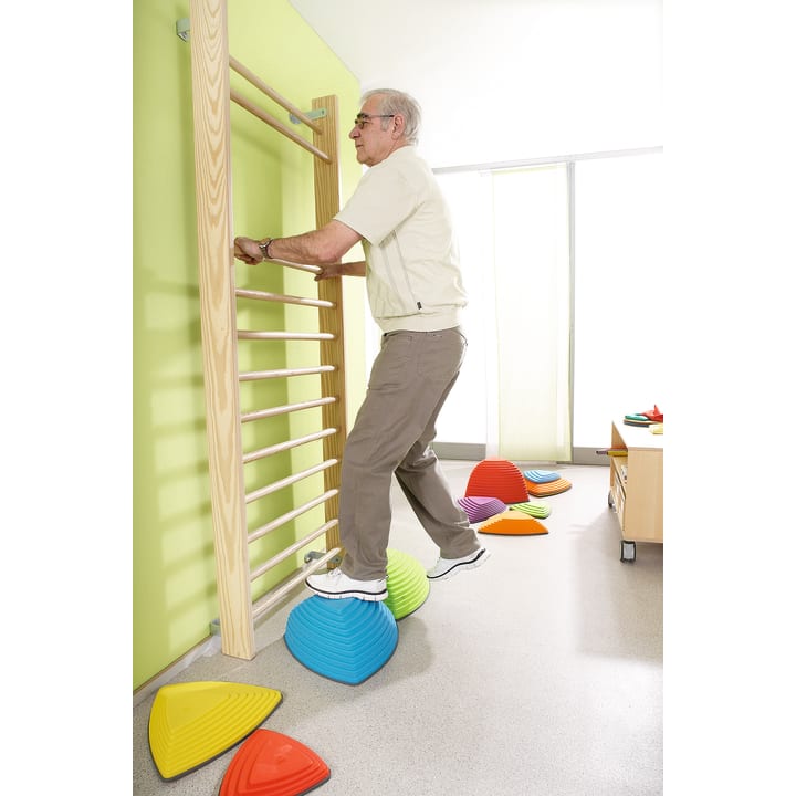 Ein älterer Mann hält sich an einer hölzernen Sprossenwand fest, während er in einem hell erleuchteten Raum auf bunte Gleichgewichtssockel auf dem Boden tritt, was auf Physiotherapie oder Gleichgewichtstraining hindeutet.