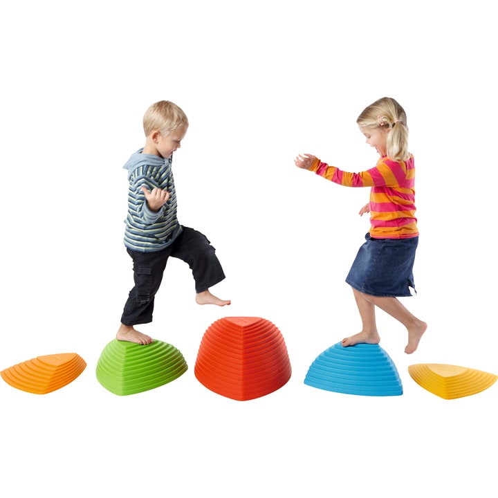 Zwei kleine Kinder balancieren auf großen, bunten, kuppelförmigen Trittsteinen, die in einer Reihe angeordnet sind. Der Hintergrund ist einfarbig weiß. Der Junge trägt einen gestreiften Kapuzenpulli und kurze Hosen, das Mädchen ein gestreiftes Oberteil und einen Jeansrock.