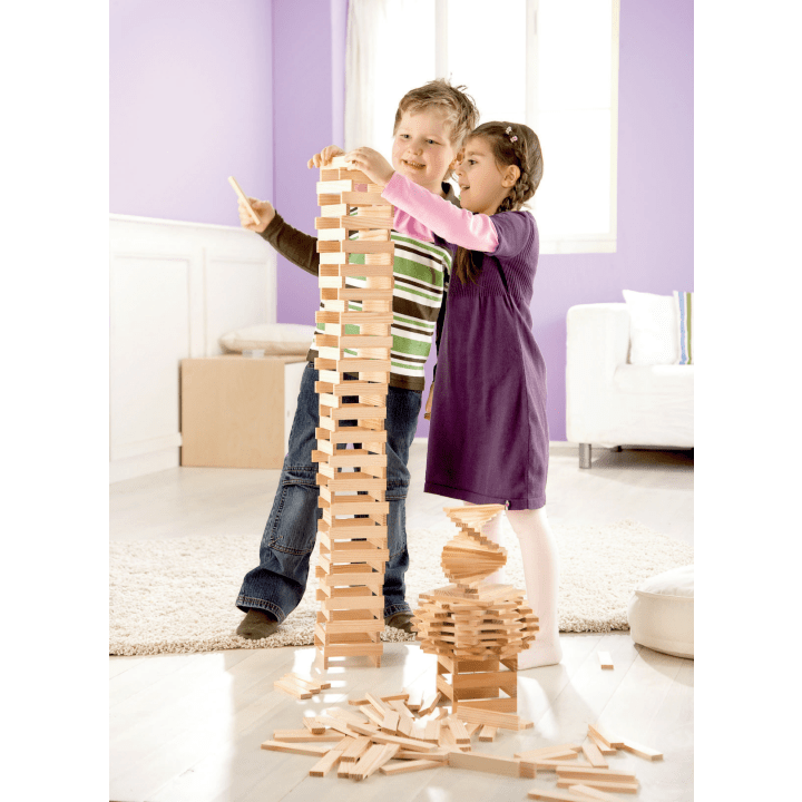 Ein Junge und ein Mädchen stehen in einem Haus und bauen einen hohen Turm aus Holzklötzen. Der Junge setzt einen Block auf die Spitze, während das Mädchen den Turm stützt. Weitere Holzklötze und ein kleineres Bauwerk liegen in der Nähe auf dem Boden.